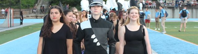 240913 DM and Guard Yorktown High School Drum Major in black and grey uniform, with white gloves and black-plumed hat and members of the Color Guard in black dance attire standing at attention in the endzone.