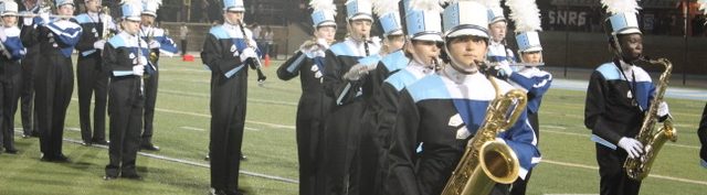 240913 Perf Bari Sax Members of the Yorktown High School Marching Band Woodwind Section performing the halftime show. Students are wearing their YHS uniforms with black pants, Navy, white, black, and light blue tops and white and black hats with white plumes.